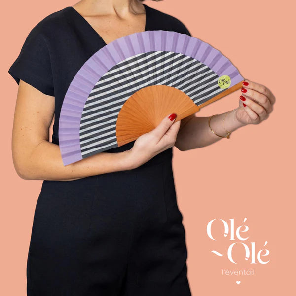 Éventail en bois fabriqué en Espagne, couleur mauve, blanc et noir, porté par une femme.